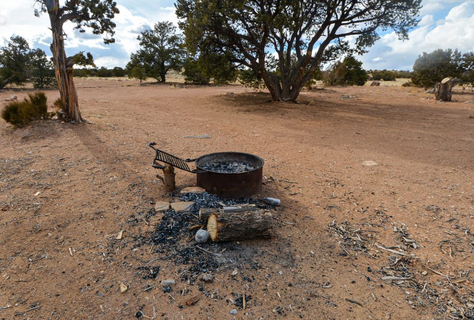 A metal campfire ring with a cooking grate at a desert campsite, with ash and a log beside it