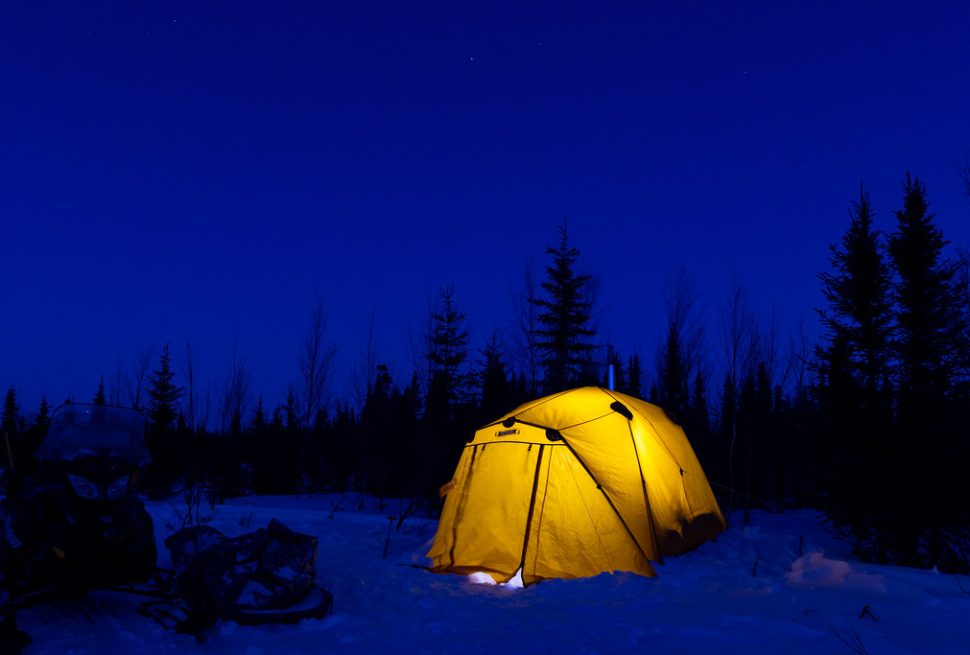 A yellow tent lit from inside on snow at dusk, with dark spruce trees behind