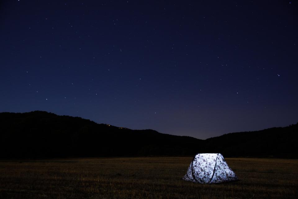 A lone tent glowing under a starry sky in an open valley below dark hills