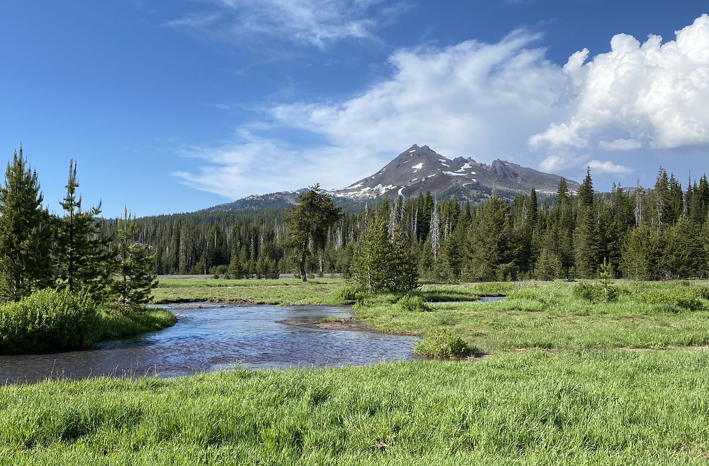 A creek winding through a green meadow below a snow-streaked mountain